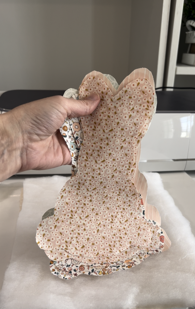 A hand holds a stack of fabric cut into the shape of a bunny, with multiple layers of pastel and floral prints. The fabric pieces are placed on top of a white batting layer, and a sewing machine is visible in the background.