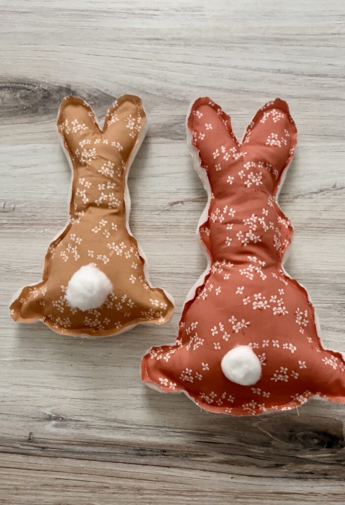 Pink and brown fabric bunnies that have cotton ball tails are shown laying on light brown wood