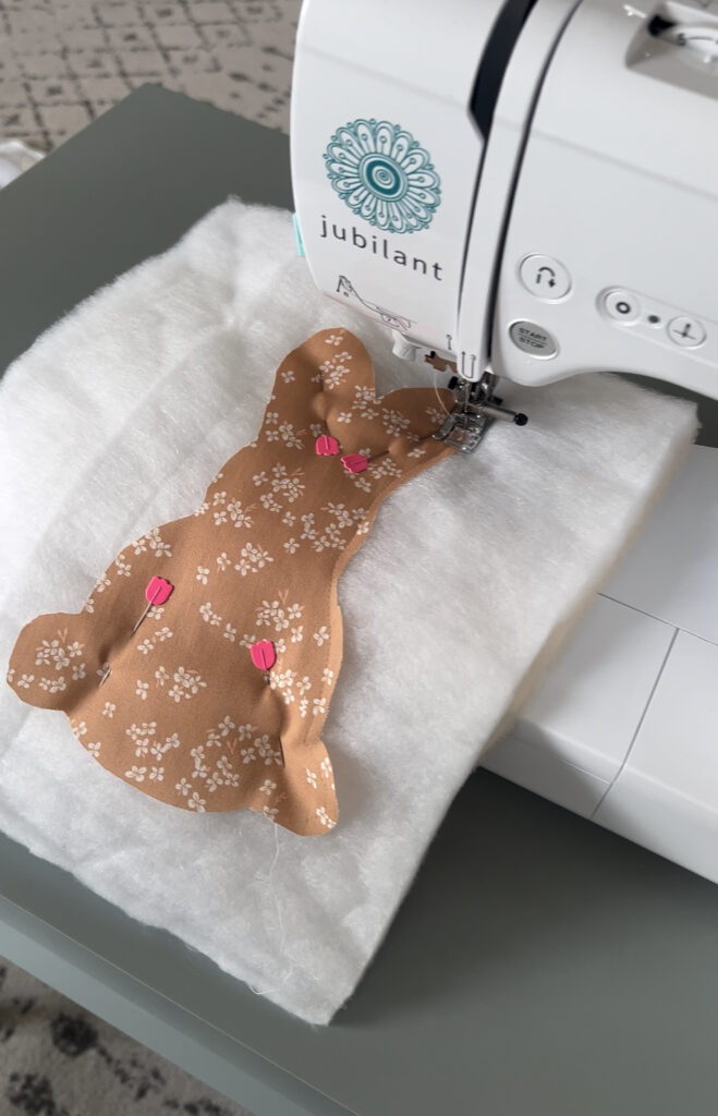 A sewing machine is stitching around a fabric bunny shape that is pinned to a layer of white batting. The fabric is a warm brown color with a delicate white floral pattern, and pink flower-shaped pins hold it in place. The sewing machine is branded "Jubilant."