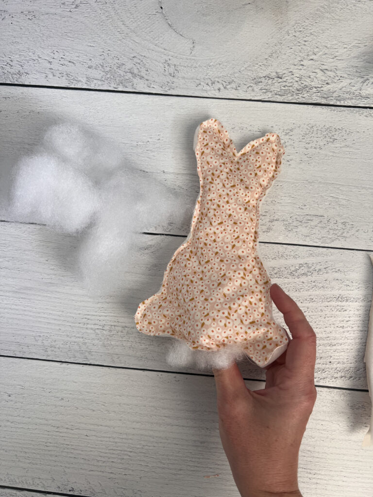 A hand holds a fabric bunny shape that has been sewn onto a layer of batting and stabilizer. The fabric features a soft pink and white floral pattern. There is cotton being used to stuff the sewn bunny.