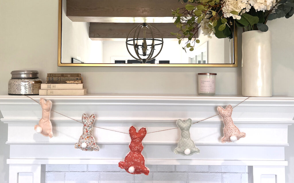 Fabric bunnies made of pink and cream fabrics are hung on a twine string decorating a white wood fireplace.