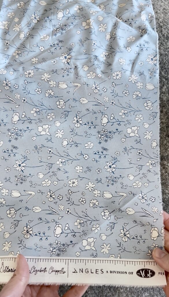 Blue fabric with small floral prints. The top of the fabric is wrinkled, the bottom is smooth. There is a hand holding the fabric at the bottom left.