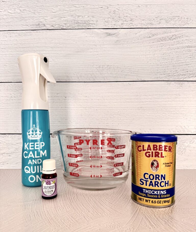 A blue spray bottle, a pyrex measuring cup, a lavender essential oil bottle, and yellow can of corn starch are placed together on a white countertop.