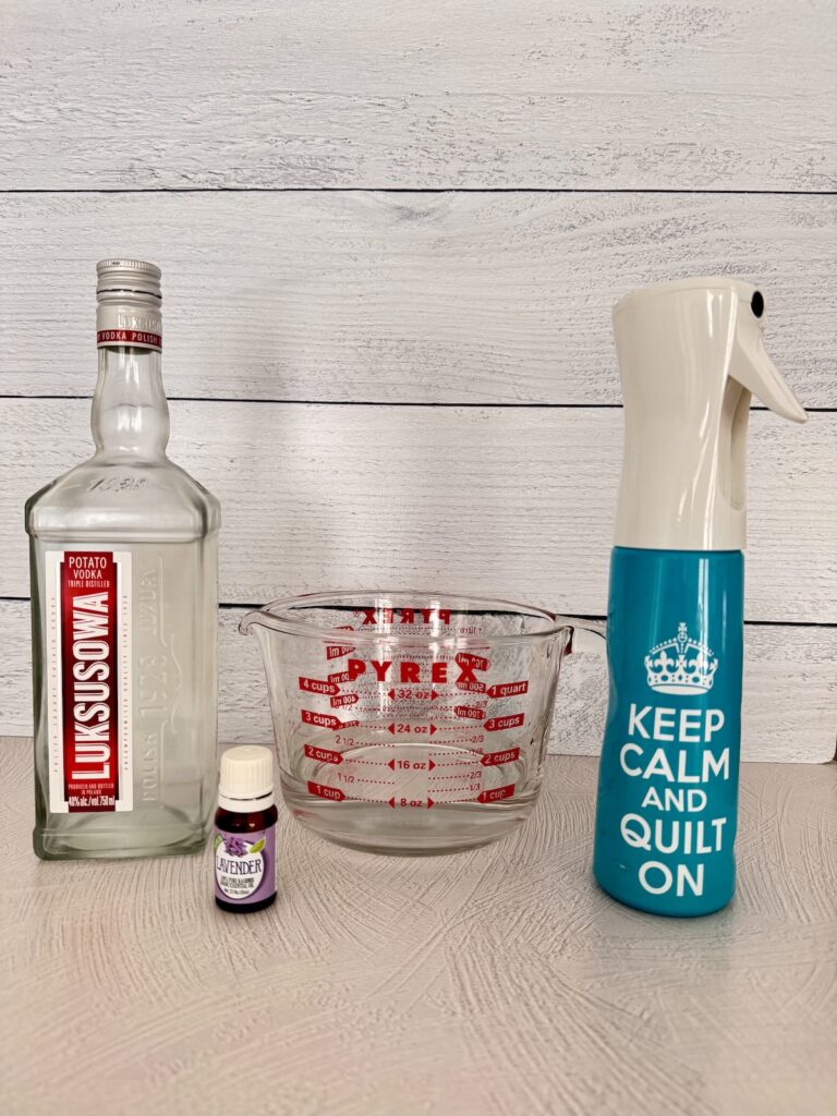 A blue spray bottle, a pyrex measuring cup, a lavender essential oil bottle, and a clear potato vodka bottle are placed together on a white countertop.