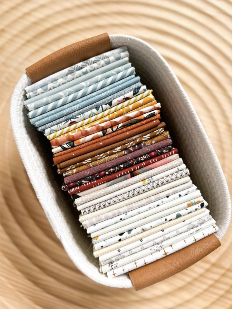 A neatly arranged basket filled with folded quilting cotton fabric in a variety of colors and patterns, including light blues, rust tones, rich burgundies, and low-volume creams and whites. The basket has leather handles and sits on a wooden surface with a circular grain pattern.