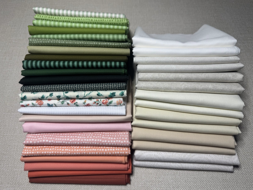 Two neatly stacked bundles of quilting fabric arranged by color. The left stack includes greens, florals, pinks, and warm coral tones, while the right stack features a gradient of whites, creams, and subtle beige prints. All fabric pieces are folded uniformly and displayed on a neutral textured surface.