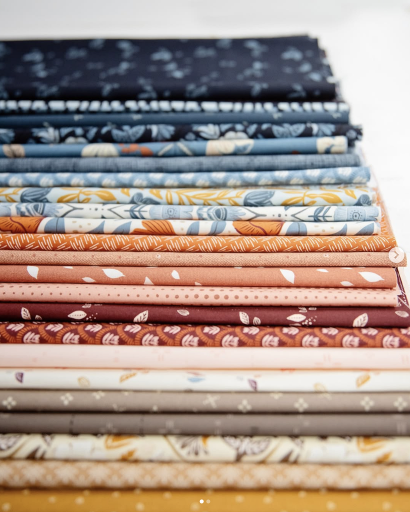 A beautifully arranged stack of folded quilting fabrics in a gradient of colors, starting with navy and denim blues at the top, transitioning through warm oranges, corals, and burgundies, and ending with soft creams, browns, and mustard yellows. Many fabrics feature botanical and leaf-inspired prints.