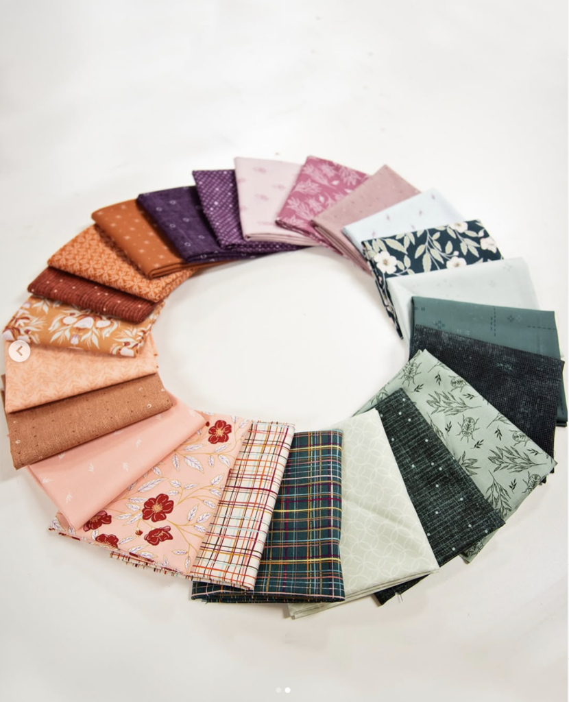 A circular arrangement of colorful quilting fabrics laid out in a gradient around a white background. The fabrics transition smoothly through shades of orange, purple, pink, floral prints, plaids, and various tones of green and teal, creating a vibrant and visually balanced color wheel.