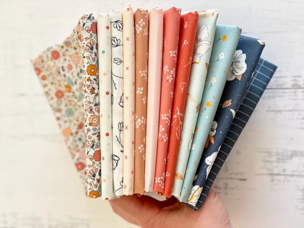 A hand holds a neatly fanned-out bundle of floral and geometric quilting cottons from the Gayle Loraine fabric collection. The palette includes soft peach, coral, cream, dusty blue, and navy tones, with charming prints of flowers, tiny dots, and linework. Perfect for quilting projects with a vintage, feminine touch.