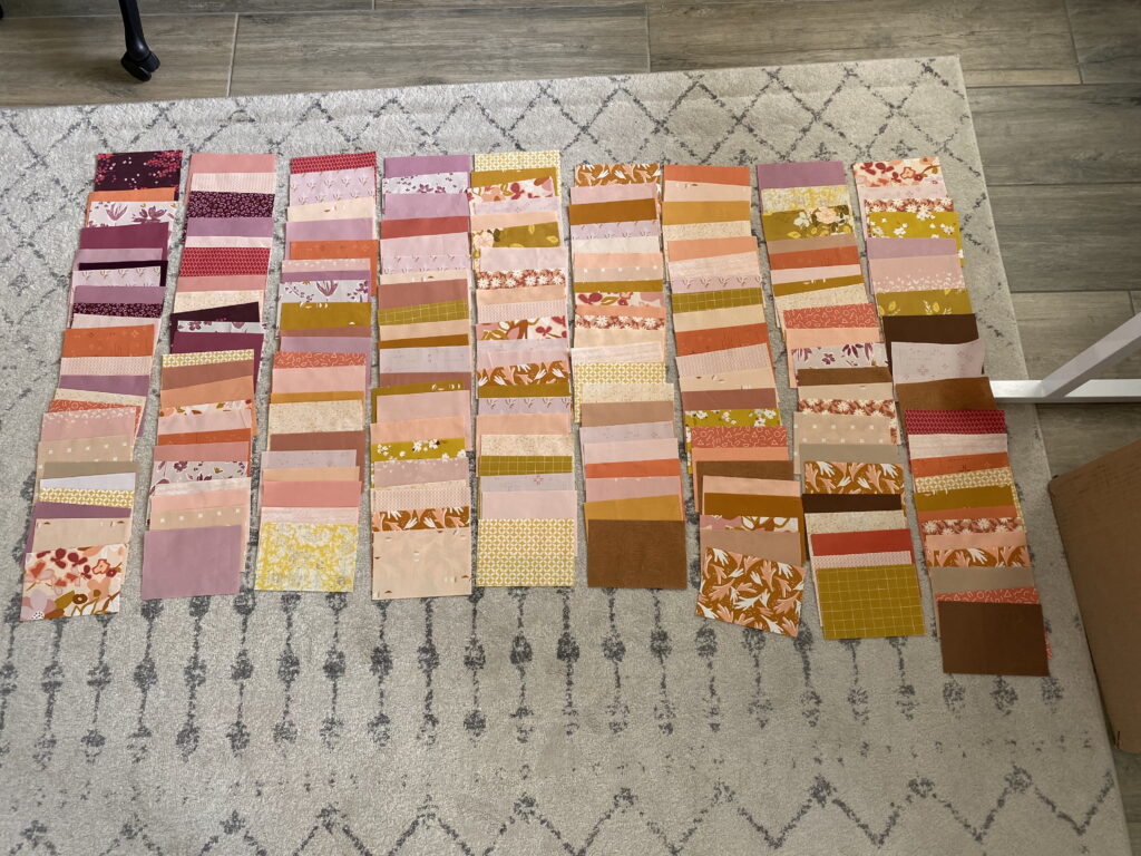 Neatly arranged quilt blocks laid out on a light patterned rug, showcasing a warm color palette of pinks, purples, golds, and oranges. The fabrics are organized in columns, helping visualize the final layout for the Front Porch Quilt.