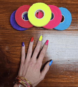 A hand with brightly painted nails in purple, red, yellow, pink, and blue rests beside rolls of Thimtec fabric thimbles in matching colors on a wooden table.