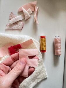 A hand holds a stitched fabric letter “F” with batting and fabric scraps on the table, demonstrating how to cut out appliqué letters after sewing.