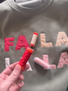 A hand holds a spool of red thread above a FA LA LA appliqué sweatshirt with matching thread colors laid out for sewing the letters.