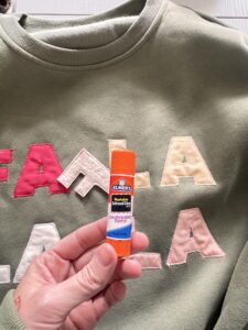 Hand holding a glue stick in front of a FA LA LA appliqué sweatshirt, showing how to secure the letters before sewing.