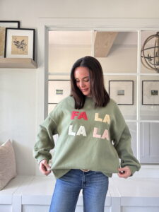 Elizabeth Chappell of Quilters Candy stands wearing a handmade green sweatshirt decorated with colorful FA LA LA appliqué letters, showing the completed project.