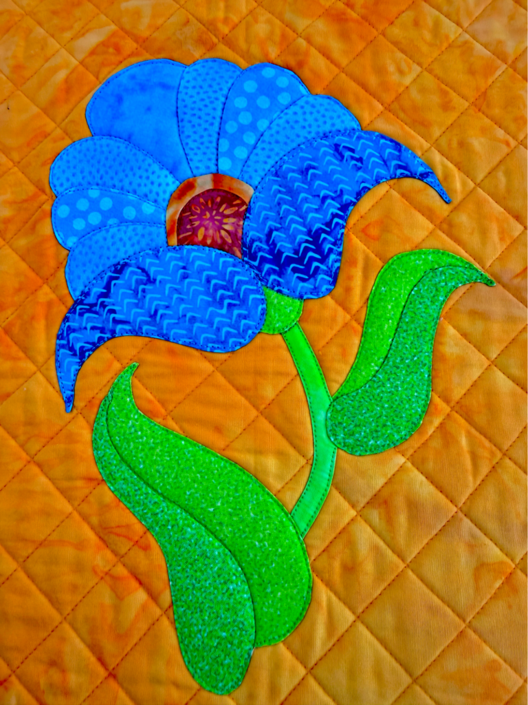 Finished turned-edge appliqué flower quilt with layered blue petals and green leaves on an orange quilted background.