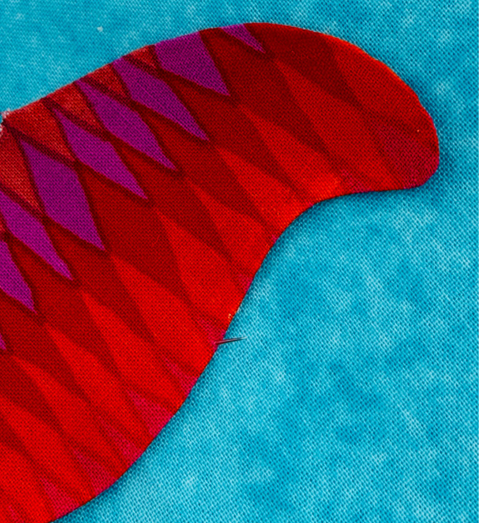 Hand needle stitching along the folded edge of a red turned-edge appliqué shape on blue fabric.