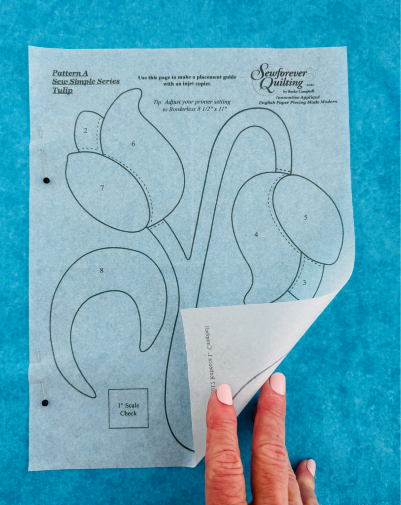 Printed appliqué placement guide pinned to blue background fabric and opened like a book for precise shape placement.
