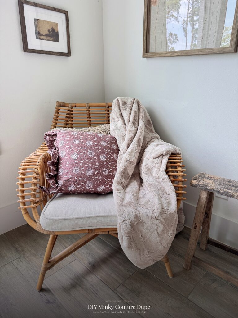Champagne colored minky couture dupe quilt draped over a rattan chair with a floral pillow