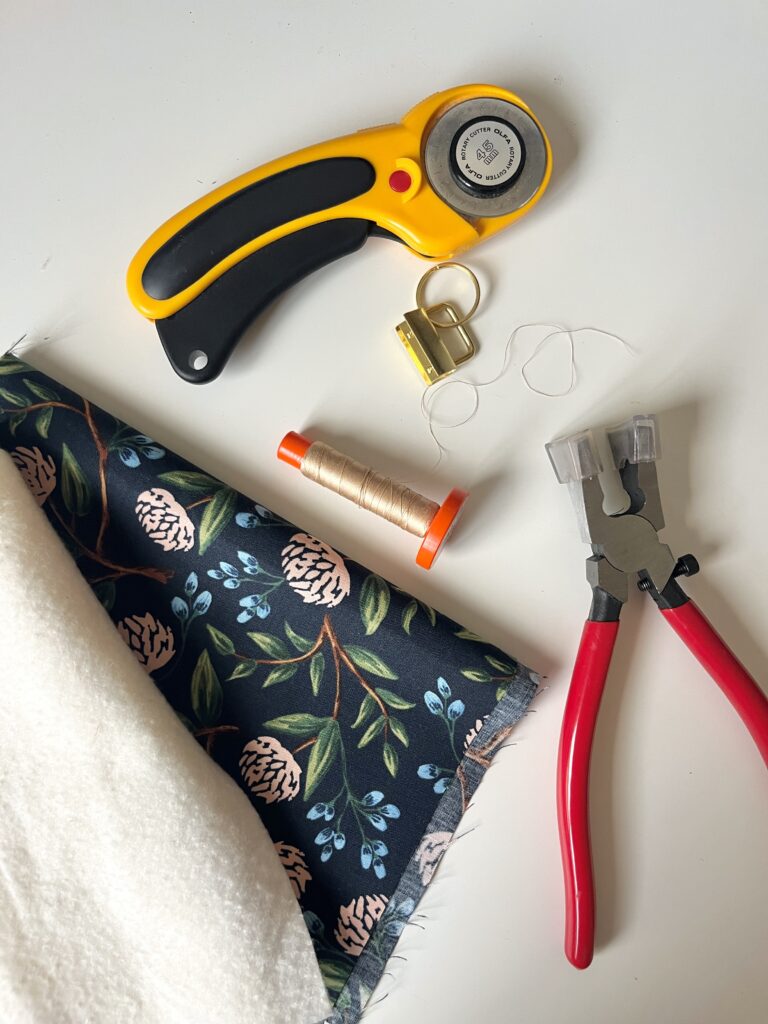 Key fob wristlet tutorial supplies: rotary cutter, gold key fob hardware, thread, floral fabric, batting, and key fob pliers