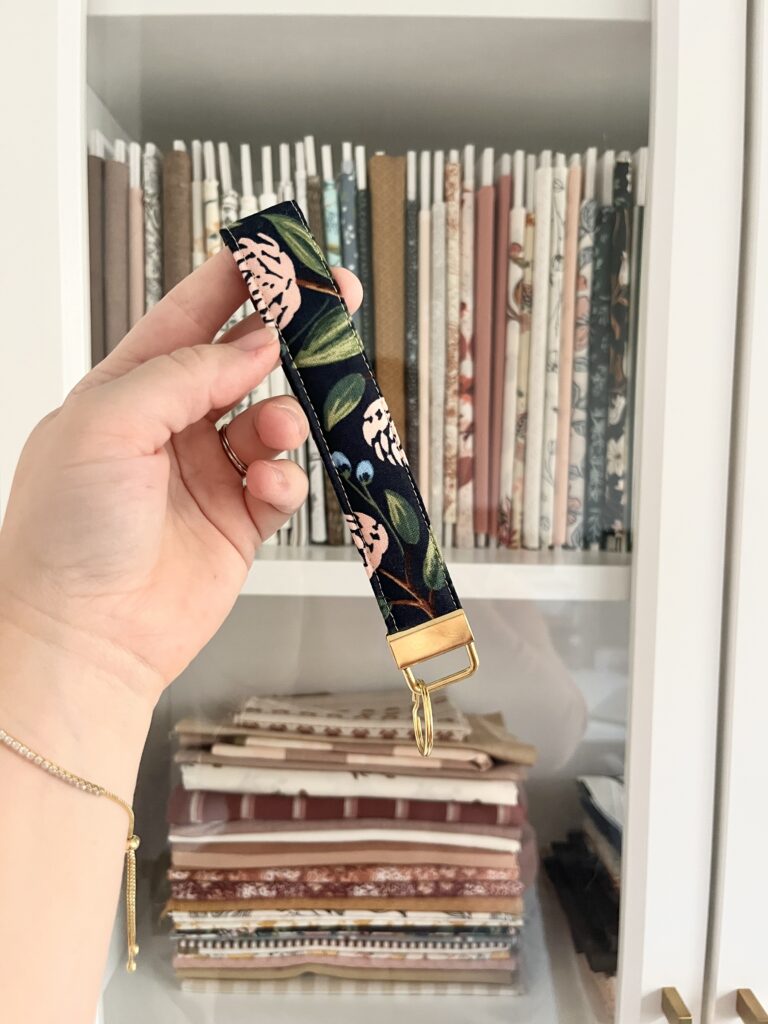 Hand holding a finished navy floral quilted key fob wristlet in front of a fabric shelf